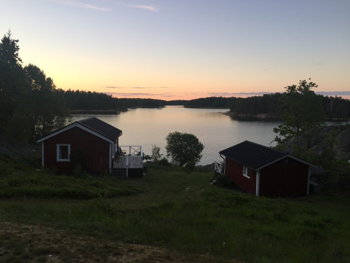 Solnedgång vid en stilla sjö med två röda stugor och en brygga, omgiven av skog.