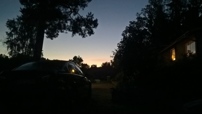 Kvällshimmel med gradient av blått till gult, silhuett av träd, en bil och ett hus med tänd lampa.