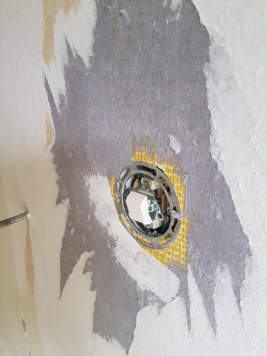 Vägg med bortskalade lager tapet och en eluttagdosa med gul vävtapet i bakgrunden.
