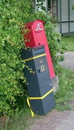 Röd och svart lutande brevlåda vid en häck, bunden med gult spännband för att matcha en granne.