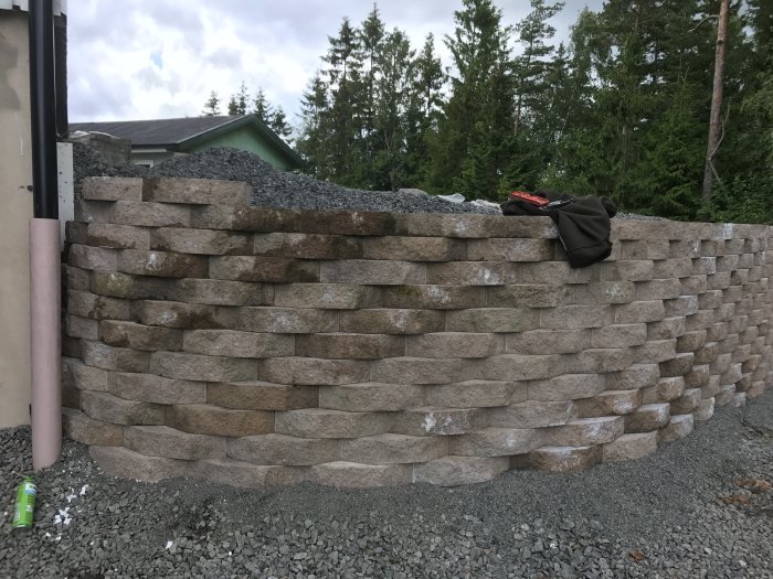 Nybyggd mur i stenblock med flera lager, en tröja ligger på toppen, grus och skog i bakgrunden.