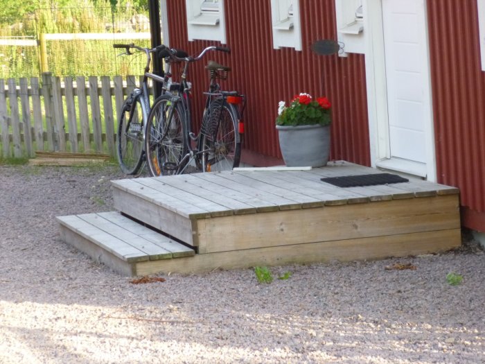Enkel trätrapp utanför en röd husfasad med cyklar, krukväxt och dörrmatta.