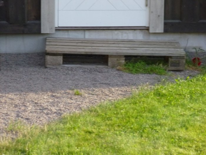 Enkel trätrapp framför en husentré omgiven av grus och gräsmatta.