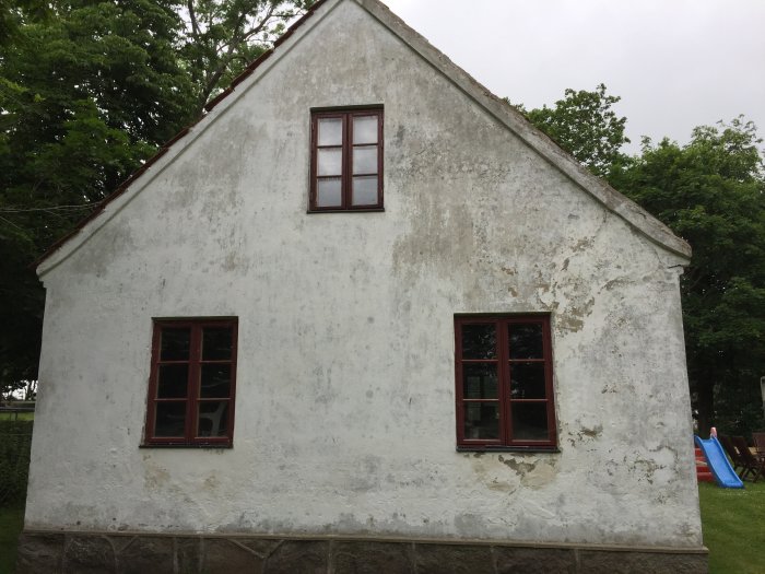 Ett äldre hus med sliten vit puts och röda fönsterkarmar, behöver renovering.