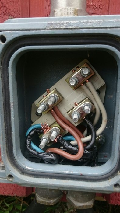Öppen elektrisk kopplingsdosa med skruvade ledningar och isoleringstejp mot röd vägg.