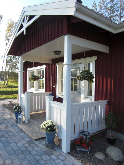 Röd husfasad med vit veranda och takpelare, prydda med blomkrukor och stenläggning framför.