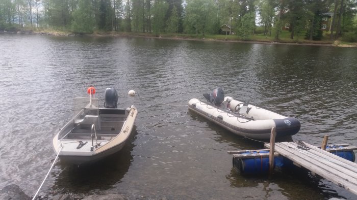 En grå gummibåt förtöjd vid en träbrygga nära en aluminiumbåt på en sjö.