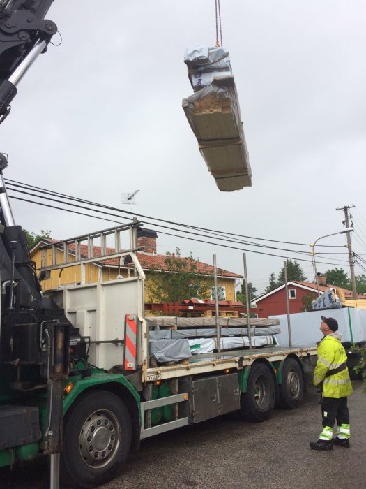 Kran lyfter byggmaterial från lastbil medan en person i skyddskläder övervakar.