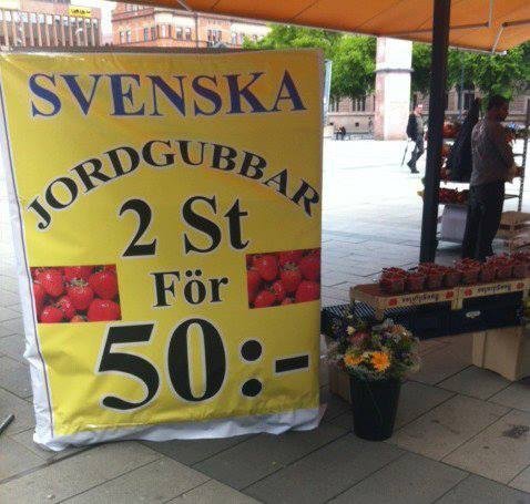 Skylt med texten "SVENSKA JORDGUBBAR 2 St För 50:-" framför en marknadsstånd med jordgubbar.