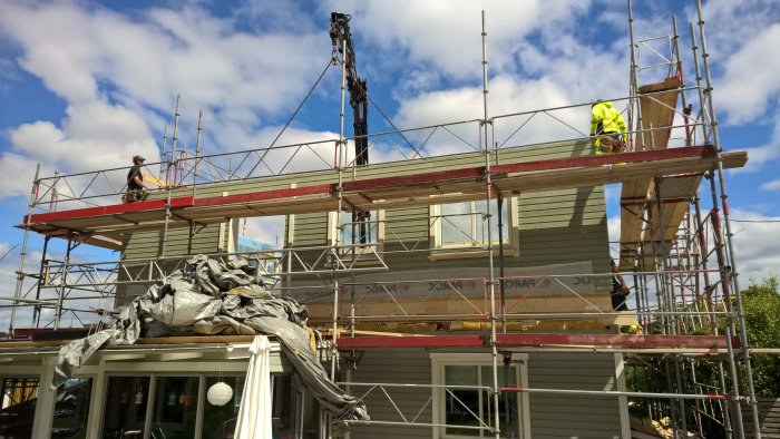 Hus under renovering med nytt bjälklag och väggelement omgivet av byggnadsställningar, arbetare synliga.