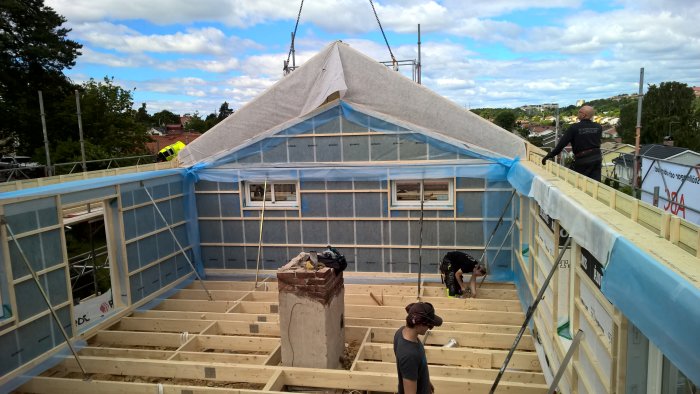 Nyinstallerat bjälklag och stående väggskelett på ett hus under konstruktion med skyddsduk på taket.