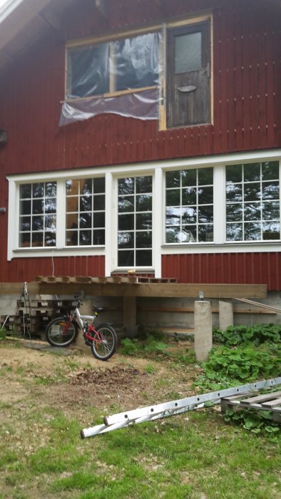 Rödfärgat hus under renovering med plasttäckt fönster och byggnadsställningar, cykel i förgrunden.