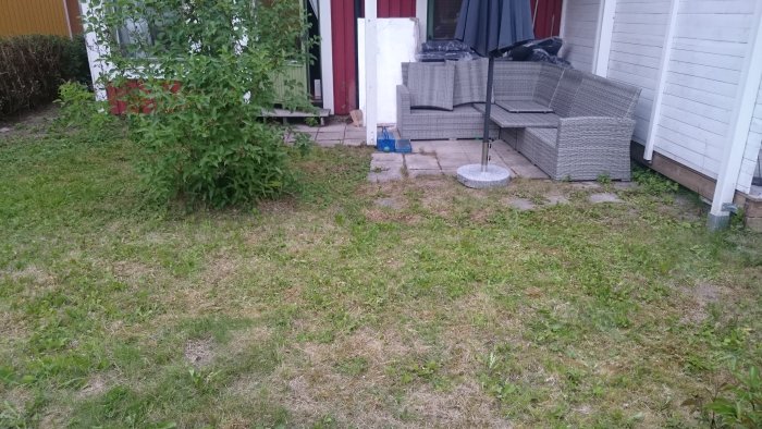 Gräsmatta framför radhus med stenplattor, trädgårdsmöbler och växter, plats för planerad veranda.