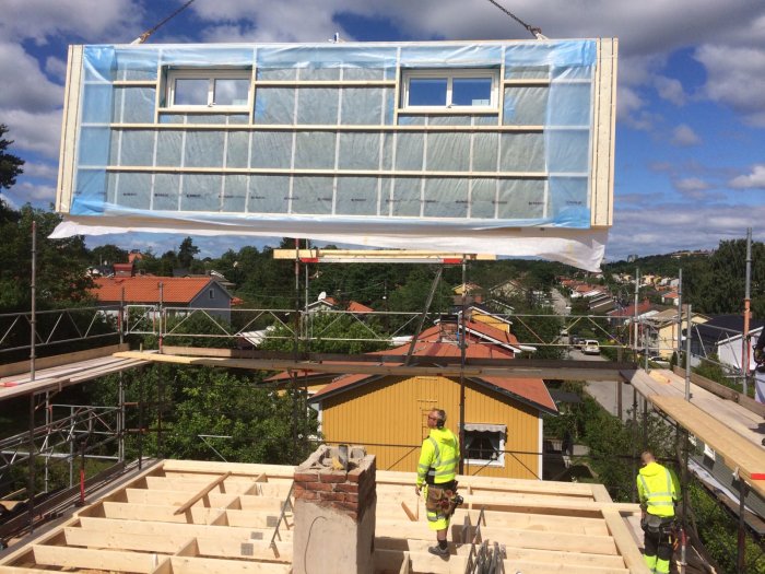 Husvåning hissas på plats av byggarbetare medan andra byggarbetare i förgrunden ser på.