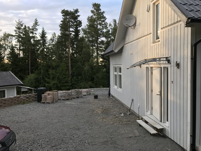 Tvåvåningshus med vit fasad och grusgång, pallar med stenplattor, skog i bakgrunden.