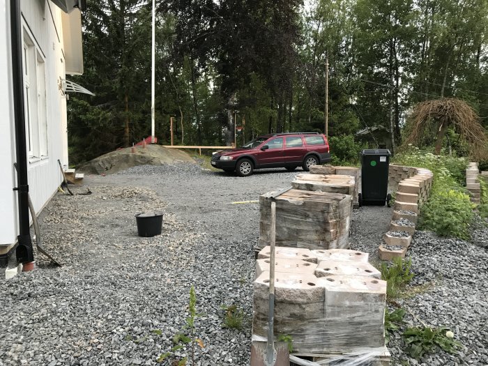 Grusad uppfart med pågående markarbete, röd bil, träpallar och grönska runtomkring.