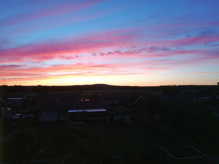 Utsikt från en taknock över bostadsområde med dramatisk solnedgång i rosa och blå toner.