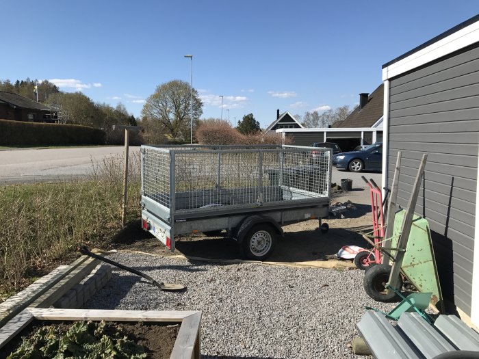 Uppgrävd rabatt intill hus och grusväg med släpkärra parkerad och grävverktyg på marken.