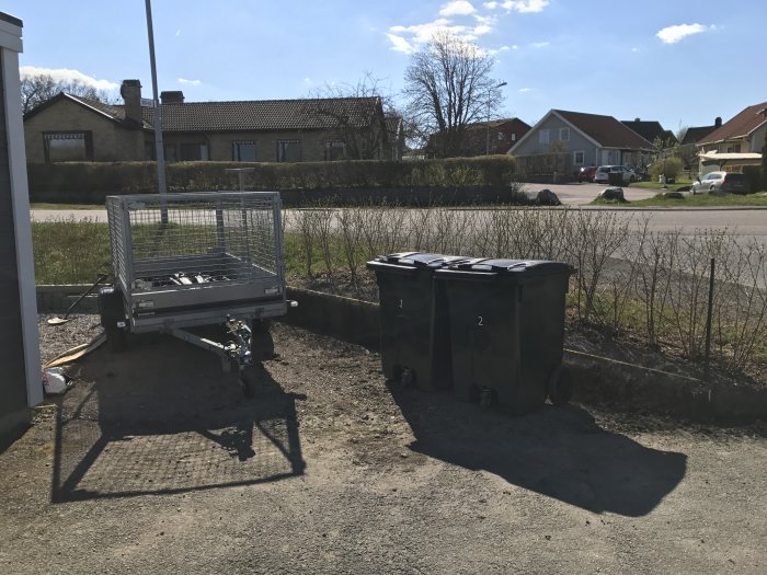 Uppställt släp och soptunnor på grusgrund där en rabatt tidigare grävts bort, med häck och hus i bakgrunden.