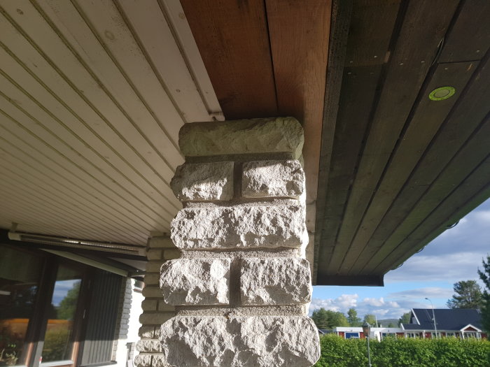 Staplad stenpelare under takutsprång med träbjälkar, intill husvägg och fönster, nära ett tak som inte möter pelaren.
