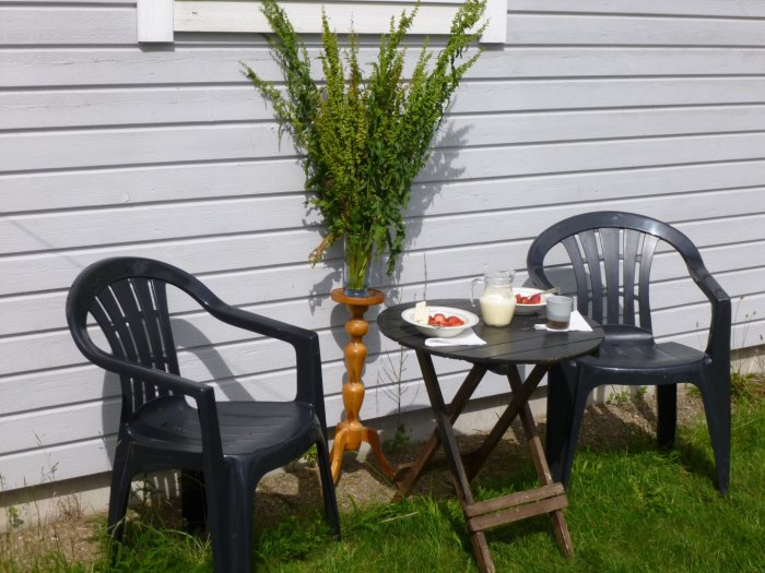Uteservering med två svarta plaststolar, träbord med fika och vas med Gårdsskräppa-blommor på träpiedestal.