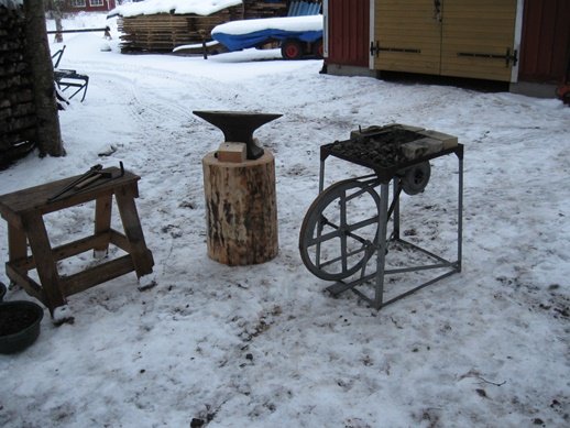 Ett traditionellt städ på en trästock och ett rälsstäd i en snöig omgivning, bredvid gamla verktyg.