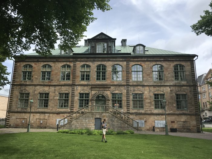 Person framför gammal byggnad med gräsmatta under en klar dag.