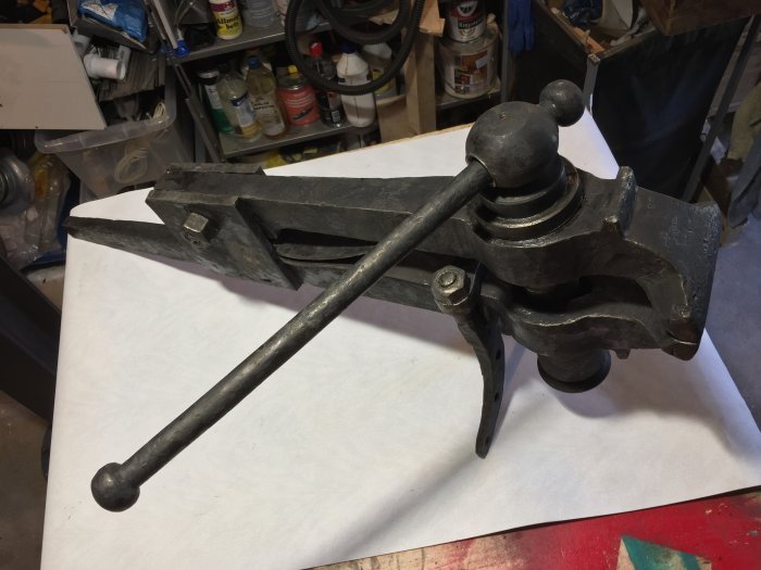 Återmonterad oxidsvart bänkskruvstäd med spänd fjäder och spak placerad på ett vitt underlag i en verkstad.