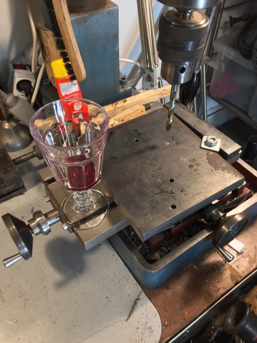 Borrpress i arbete med stadig metallplatta, närbild på borrtopp, och ett glas Zinfandel på arbetsbänken.