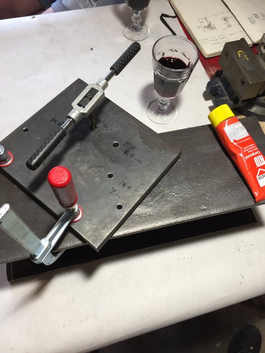 Metallstativ i konstruktion med borr, gängtapp och smörjmedel på arbetsbänk, bredvid ett glas rött vin.