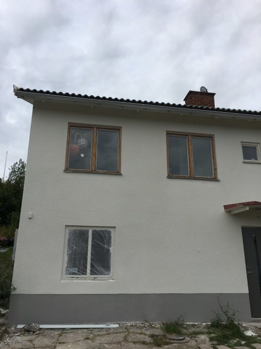 Nyputsad fasad på hus med oputsade fönsterkarmar och en skyddad fönsterruta i grått väder.