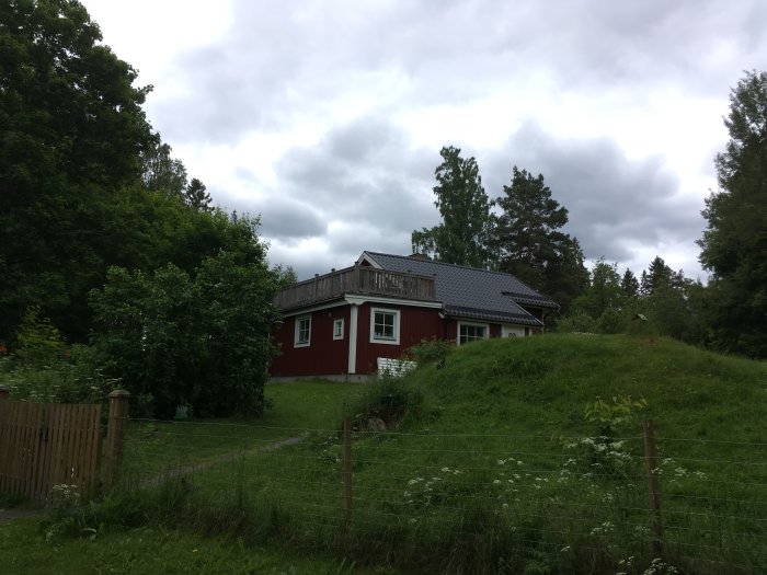 Rött hus med takterrass och staket i en grönskande trädgård, molnig himmel i bakgrunden.