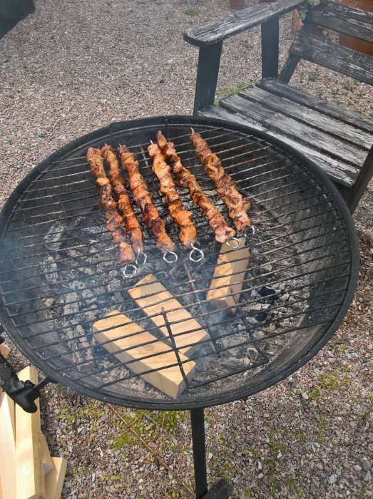 Grillning av köttspett över ved på en rund kolgrill utomhus nära slitna trädgårdsbänkar.