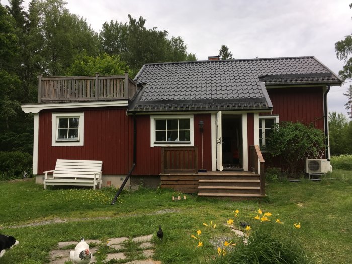 Röd trävilla med svart tak och veranda, omgiven av grönska och några höns framför huset.