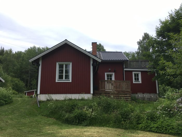 Röd stuga med vita knutar och en framträdande skorsten, omgiven av grönt gräs och träd, med en träveranda.