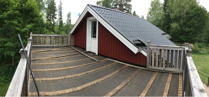 Panoramabild av en svängd träaltan med räcke framför en rödfärgad stuga med grått tak.