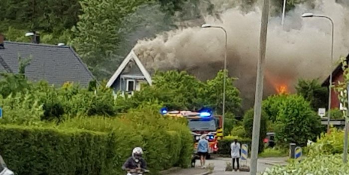 Brandbil med blåljus framför kraftig rök och eld från husbrand, omgiven av grönska.