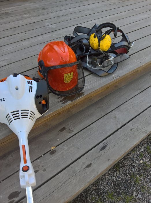 Skogshjälm med visir och hörselskydd vid sidan av en STIHL röjsåg på ett trägolv utomhus.