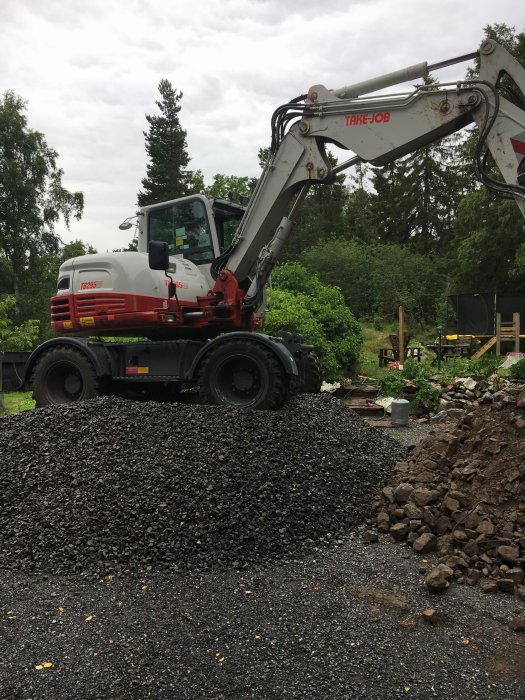 Grävmaskin bredvid hög med grus och schaktmassor på en byggarbetsplats med grönska i bakgrunden.