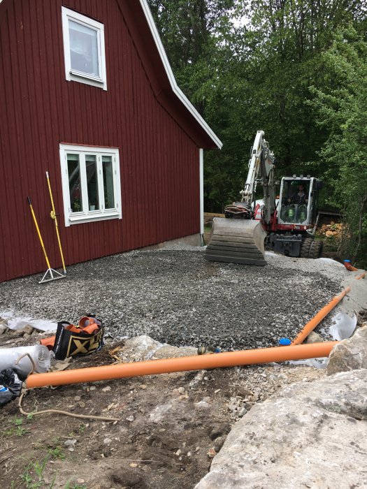 Grundarbete vid hus med grus och nyinstallerade dagvattenrör, grävmaskin i bakgrunden och byggmaterial synligt.