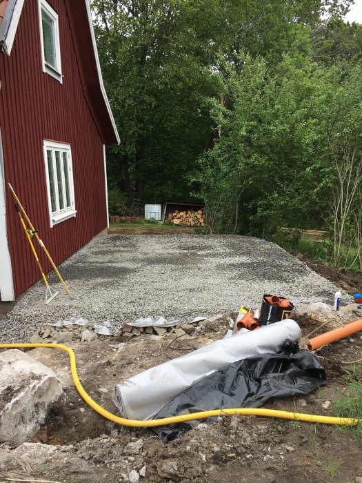 Grundarbete för hus med grus, dagvattenledning och verktyg nära röd byggnad.