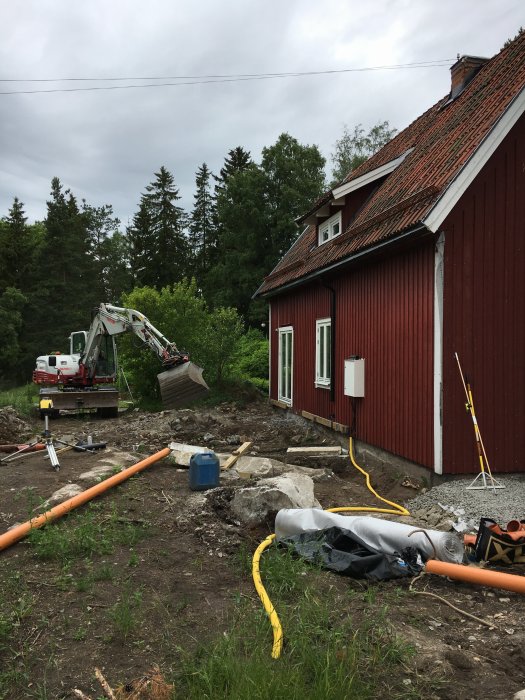 Grundarbete vid röd huskropp med grävmaskin, orange dagvattenrör och byggmaterial på marken.