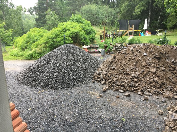 Två grushögar för byggprojekt, en av finmakadam och en av större sten, i en trädgård med lekställning i bakgrunden.