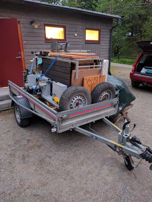 Släpvagn fullastad med verktyg och möbler, däribland en byggsåg och snickarbänk, framför en stuga.