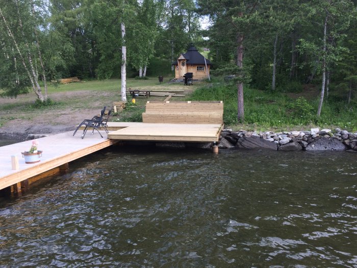 Nybyggd träbrygga vid en sjö med stolar och en småstuga i bakgrunden omgiven av träd.