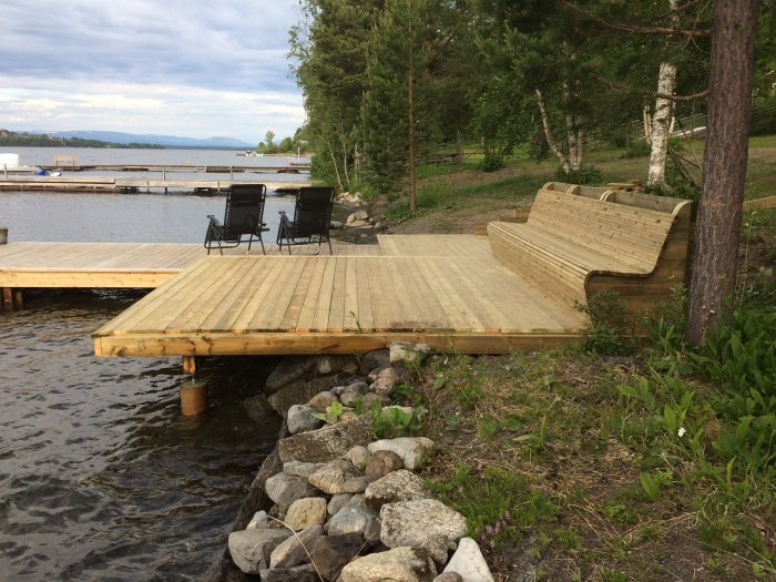 Träbrygga med inbyggda sittplatser vid en sjö, omgiven av stenar och grönska med en flytbrygga i bakgrunden.