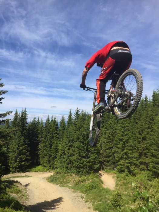 Cyklist i rött utför ett hopp med mountainbike mot skogsbakgrund under blå himmel.