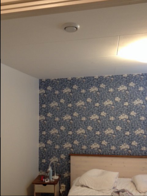 En tilluftsventil i taket över en säng med blått blommigt tapetserad vägg i ett sovrum.