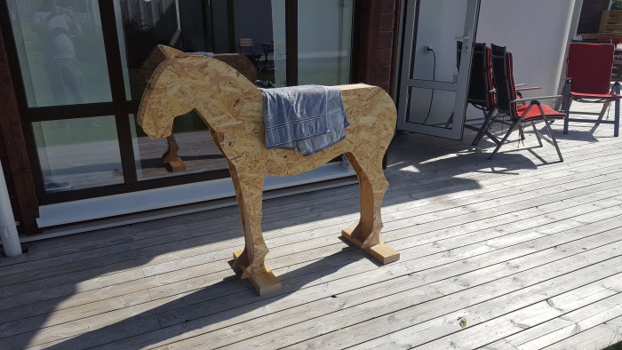 Hemmagjord lekshäst av OSB-skivor på en träveranda med en handduk över ryggen.