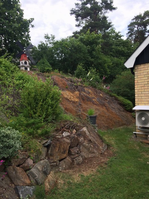 Brant tomt med berg och ojämna ytor vid husets baksida, omgiven av vegetation.